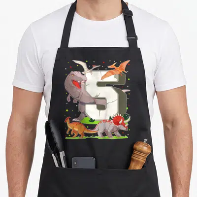 Tablier de cuisine noir avec un grand chiffre 5 blanc fissuré entouré de dinosaures colorés et d'étoiles multicolores sur fond gris uni.