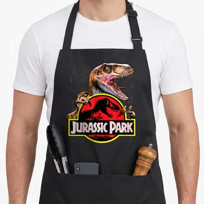 Tablier de cuisine noir avec un raptor au-dessus du logo Jurassic Park rouge et jaune.