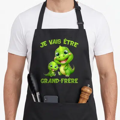 Tablier de cuisine noir avec deux dinosaures verts souriants et le texte Je vais être grand-frère imprimé en vert clair.