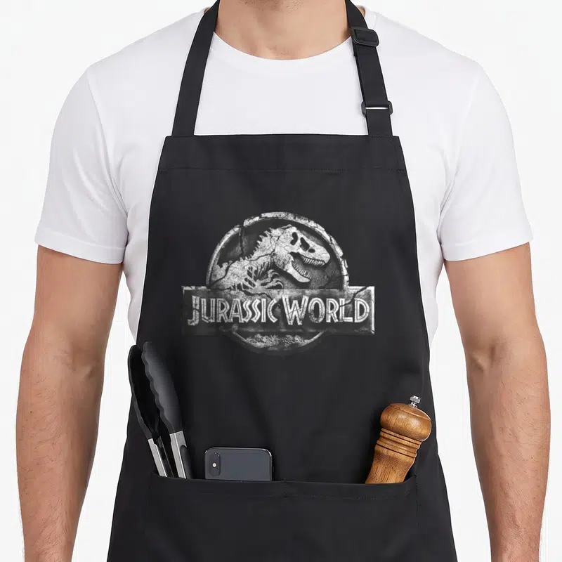 Tablier de cuisine noir avec logo Jurassic World représentant un dinosaure crachant en relief pierre fissurée.