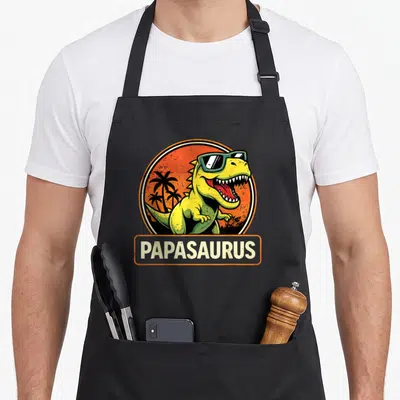 Tablier de cuisine noir avec un t-rex jaune portant des lunettes vertes et le mot Papasaurus sur fond orange.
