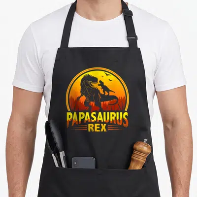 Tablier de cuisine noir avec illustration de dinosaures et texte Papasaurus Rex en orange.