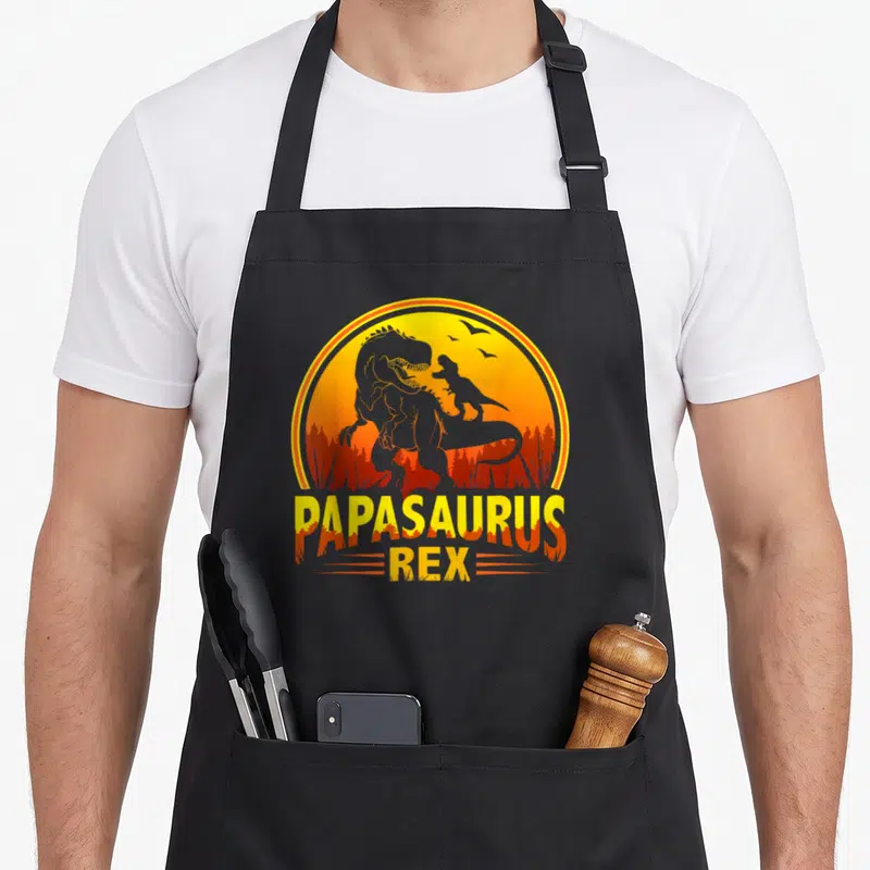 Tablier de cuisine noir avec illustration de dinosaures et texte Papasaurus Rex en orange.