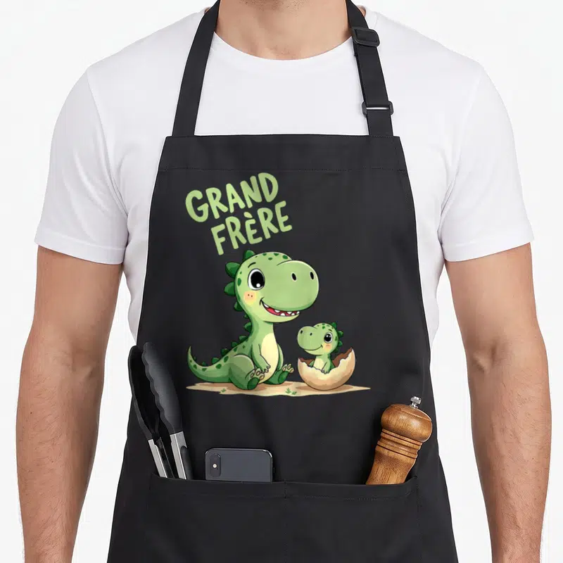 Tablier de cuisine noir avec illustration d'un dinosaure vert souriant et d'un bébé dinosaure dans une coquille d'œuf brisée.