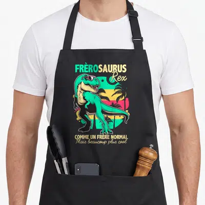 Tablier de cuisine noir avec illustration colorée d'un dinosaure vert portant des lunettes de soleil roses et du texte humoristique.