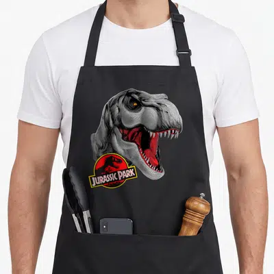 Tablier de cuisine noir avec tête de T-Rex en pointillisme et logo Jurassic Park rouge et jaune en bas à gauche.