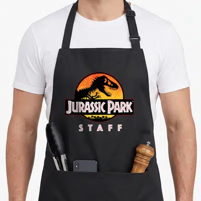 Tablier de cuisine noir avec le logo Jurassic Park Staff représentant un T-Rex sur fond de soleil orange.