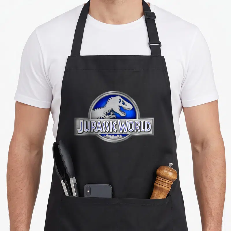 Tablier de cuisine noir avec logo circulaire bleu montrant un squelette de dinosaure et l'inscription Jurassic World en lettres argentées.