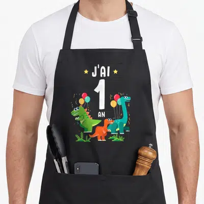 Tablier de cuisine noir avec trois dinosaures colorés tenant des ballons et le texte J'ai 1 an au centre.