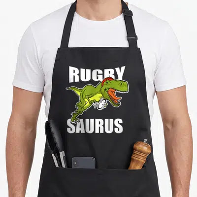 Tablier de cuisine noir avec illustration d'un dinosaure vert tenant une balle de rugby sous le mot Rugby Saurus