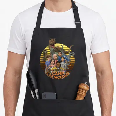 Tablier de cuisine noir avec illustration colorée du groupe Camp Cretaceous et dinosaures sur fond circulaire jaune rayé.
