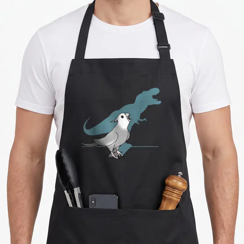 Tablier de cuisine noir avec illustration d'une calopsitte blanche projetant l'ombre bleue d'un dinosaure T-Rex.