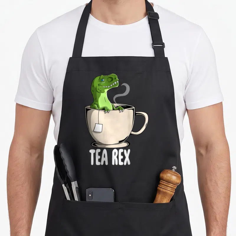 Tablier de cuisine noir avec un Tyrannosaure vert assis dans une tasse de thé fumante sous l'inscription Tea Rex.