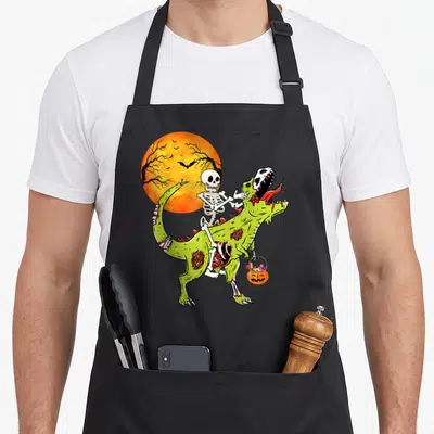 Tablier de cuisine noir avec illustration d'un squelette montant un dinosaure vert zombie sous une lune orange d'Halloween.