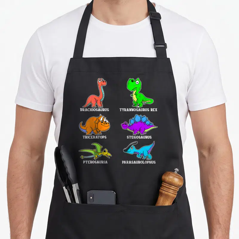 Tablier de cuisine noir avec six dinosaures colorés et leurs noms imprimés en blanc sous chaque illustration.