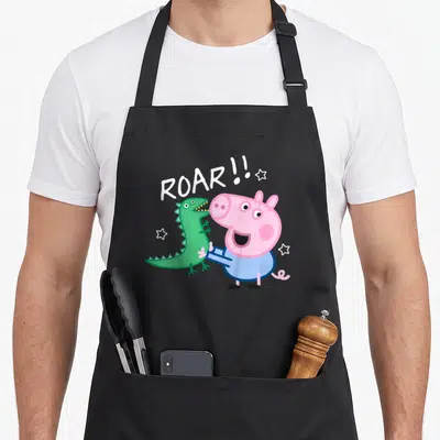 Tablier de cuisine noir avec George en costume de dinosaure vert et l'inscription ROAR entourée d'étoiles blanches sur fond gris uni.