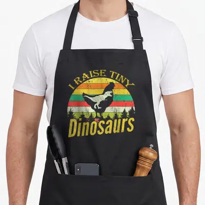 Tablier de cuisine noir avec un motif vintage montrant un poulet en forme de dinosaure sur fond arc-en-ciel et texte jaune.