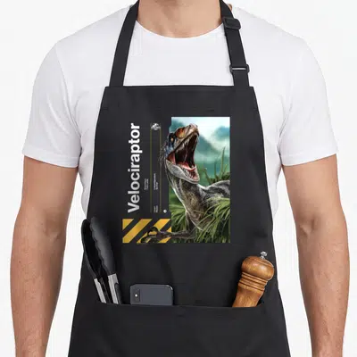 Tablier de cuisine noir avec illustration réaliste d'un Velociraptor gueulant et bandes jaunes de signalisation en bas à gauche.