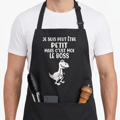 Tablier de cuisine noir avec un dessin de T-Rex blanc et le texte Je suis petit mais c'est moi le boss imprimé en haut.