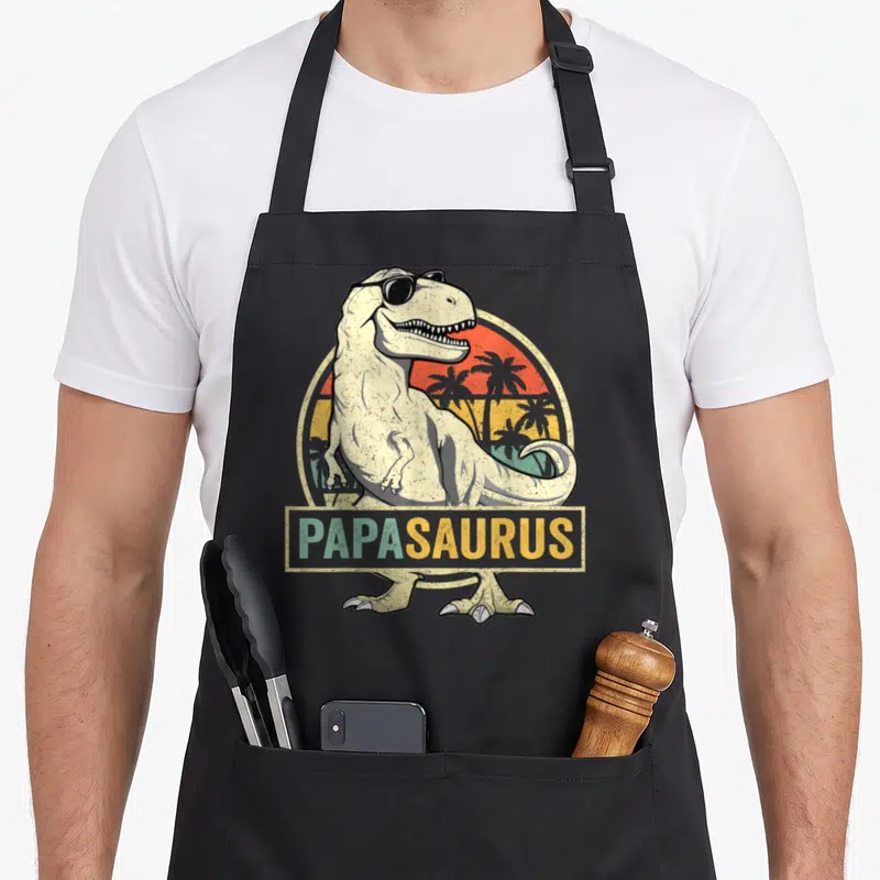Tablier de cuisine noir avec un Tyrannosaurus Rex en lunettes de soleil et le mot Papa Saurus sur fond de soleil tropical.