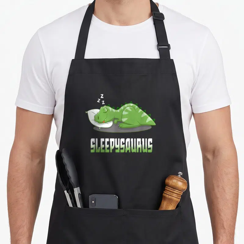 Tablier de cuisine noir avec un dinosaure vert dormant sur un coussin blanc et le texte Sleepysaurus