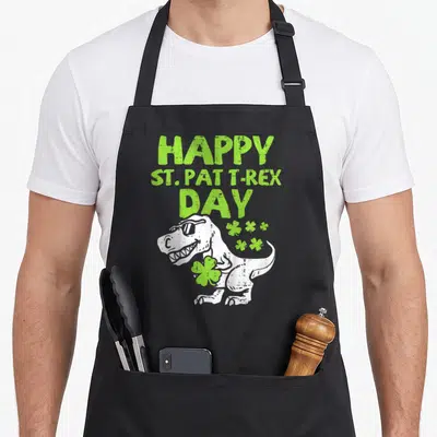 Tablier de cuisine noir avec Tyrannosaurus aux lunettes de soleil tenant un trèfle vert et le texte Happy St Pat T-Rex Day en vert.