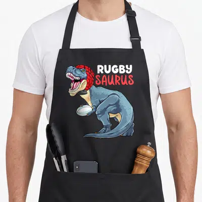Tablier de cuisine noir avec illustration d'un dinosaure bleu tenant un ballon de rugby sous le texte Rugby Saurus.