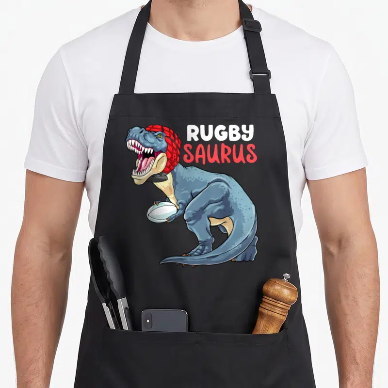Tablier de cuisine noir avec illustration d'un dinosaure bleu tenant un ballon de rugby sous le texte Rugby Saurus.