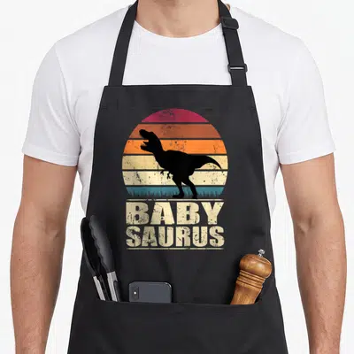 Tablier de cuisine noir avec silhouette T-Rex et texte Baby Saurus sur fond de soleil rétro multicolore.