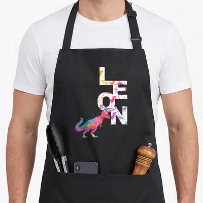 Tablier de cuisine noir avec le nom Leon en lettres pixelisées blanches et un dinosaure multicolore au style art numérique.