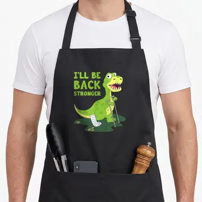 Tablier de cuisine noir avec dinosaure vert sur béquilles et texte I'll be back stronger imprimé en lettres capitales vertes.