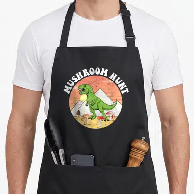 Tablier de cuisine noir avec illustration d'un dinosaure vert cherchant des champignons devant des montagnes.