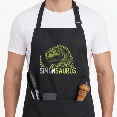 Tablier de cuisine noir avec illustration verte d'un dinosaure Tyrannosaurus Rex et le texte Simon Saurus imprimé en blanc et vert.