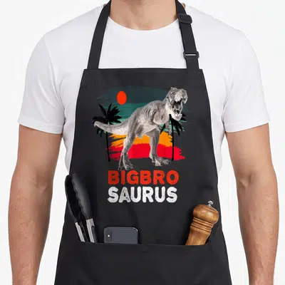 Tablier de cuisine noir avec illustration d'un T Rex sous des palmiers et le texte Bigbro Saurus en lettres rouges et blanches.