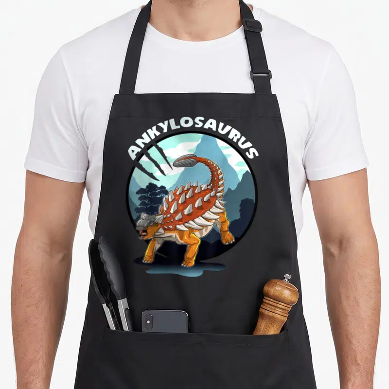Tablier de cuisine noir avec illustration d'un Ankylosaurus orange et blanc dans un cadre circulaire bleu et gris.