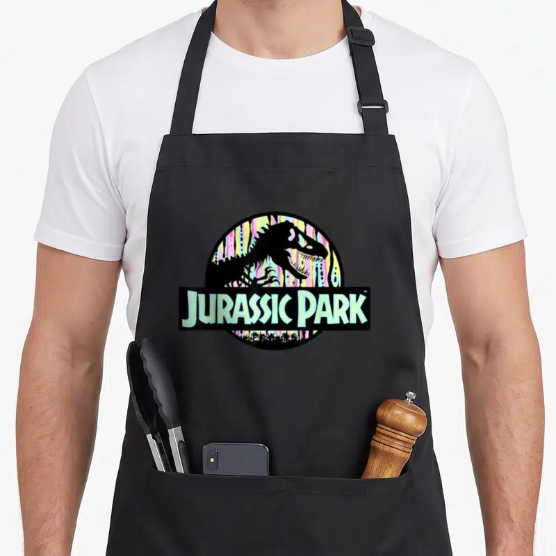 Tablier de cuisine noir avec logo Jurassic Park aux couleurs pastel et silhouette de dinosaure.