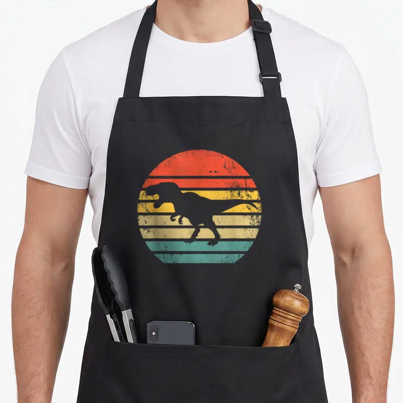 Tablier de cuisine noir avec silhouette T-rex grise sur fond de soleil rétro aux bandes orange jaune et turquoise striées.