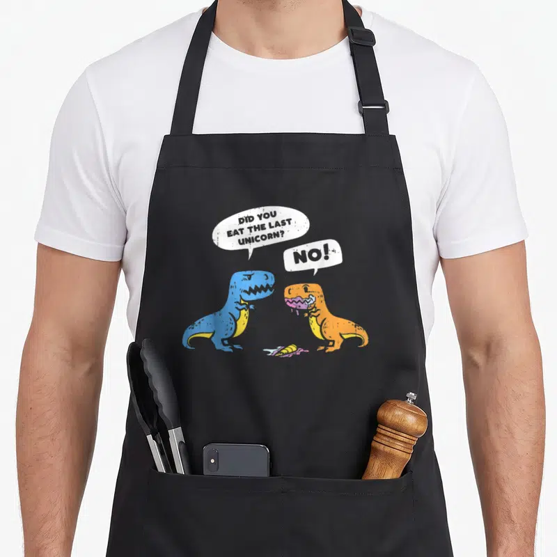 Tablier de cuisine noir avec deux dinosaures colorés discutant de la dernière licorne mangée.