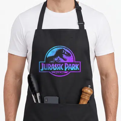 Tablier de cuisine noir avec logo Jurassic Park en dégradé bleu et violet sur fond gris uni.