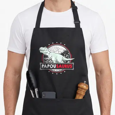 Tablier de cuisine noir avec illustration d'un Tyrannosaure vert mint et le mot PapouSaurus en blanc et rouge.