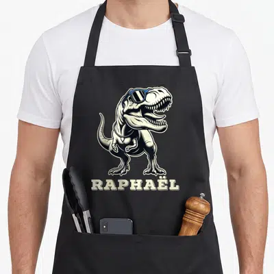 Tablier de cuisine noir avec illustration d'un dinosaure aux lunettes bleues et le nom Raphaël imprimé en bas.