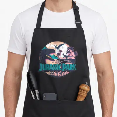 Tablier de cuisine noir avec illustration circulaire montrant un dinosaure, des palmiers et le texte Jurassic Park en lettres turquoise.