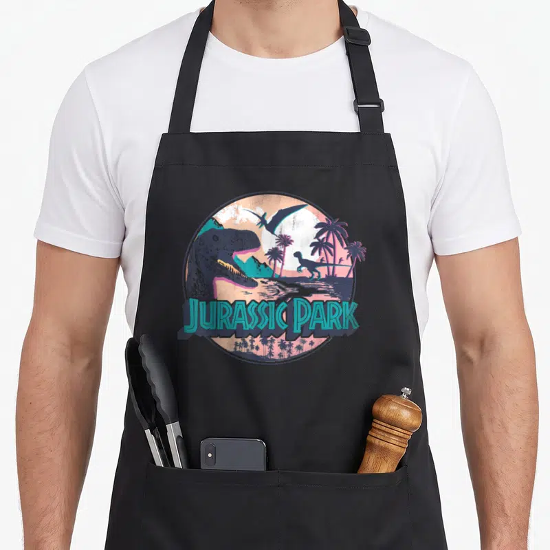 Tablier de cuisine noir avec illustration circulaire montrant un dinosaure, des palmiers et le texte Jurassic Park en lettres turquoise.