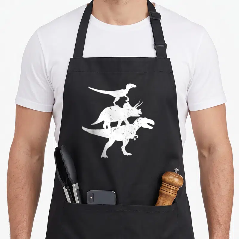 Tablier de cuisine noir avec silhouette blanche de trois dinosaures superposés dont un Raptor et un T-Rex.