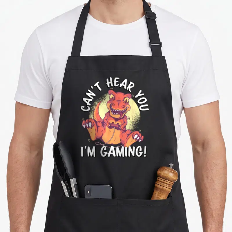 Tablier de cuisine noir avec un T-Rex orange tenant une manette et le texte Can't Hear You I'm Gaming.