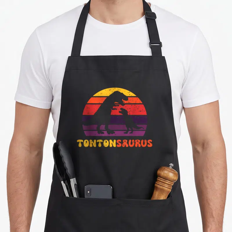 Tablier de cuisine noir avec silhouette de dinosaures et inscription Tontonsaurus en lettres jaunes et oranges.