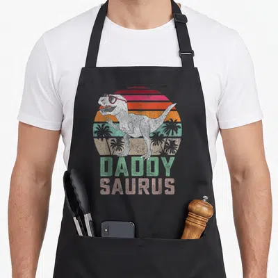 Tablier de cuisine noir avec illustration d'un T Rex portant des lunettes de soleil et le texte Daddy Saurus en lettres colorées.