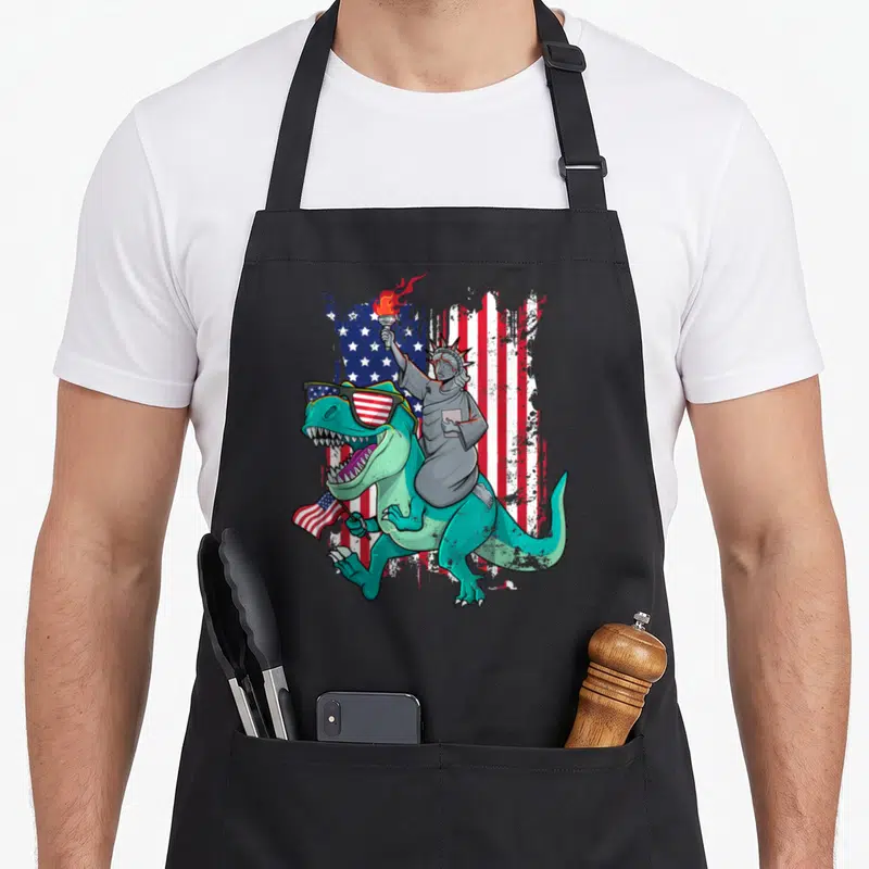 Tablier de cuisine noir avec Statue de la Liberté chevauchant un T-Rex aux lunettes à drapeau américain.