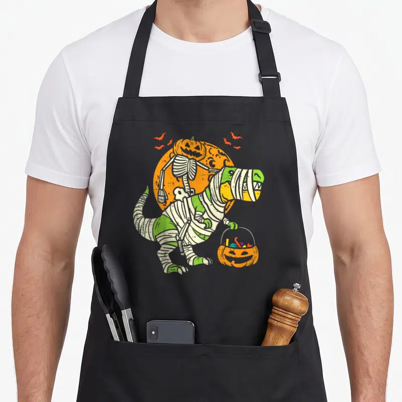 Tablier de cuisine noir avec illustration T-Rex mummy et squelette citrouille sur fond orange.