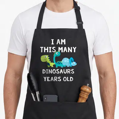 Tablier de cuisine noir avec trois dinosaures mignons et le texte Je suis ce nombre de dinosaures trois ans en blanc.